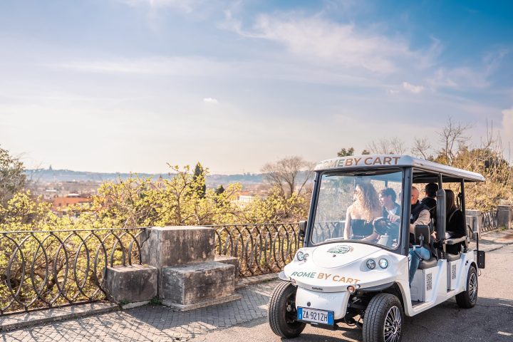 A small white cart with people inside driving along a scenic road with trees and blue sky in the background.