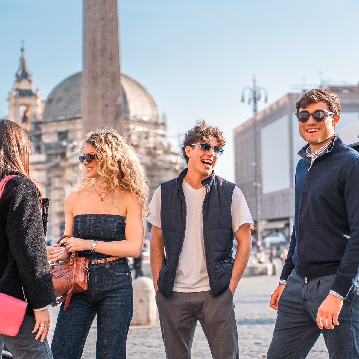 Four people in sunglasses laughing and talking in a sunny urban setting with historical buildings.