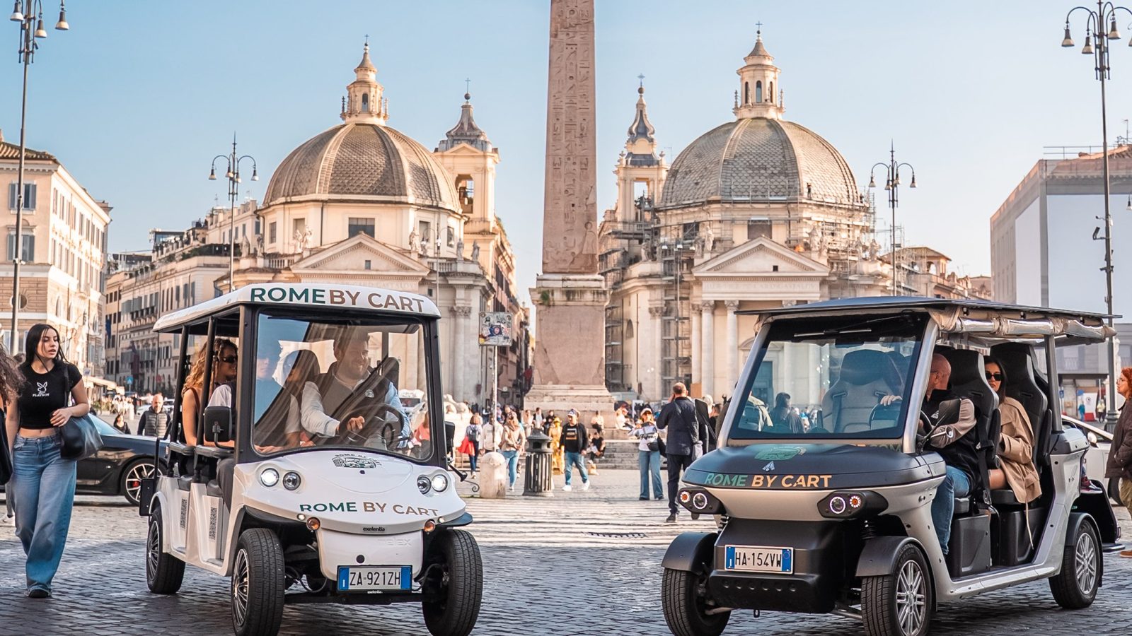 Two golf carts on a cobblestone street with domed buildings and an obelisk in the background.