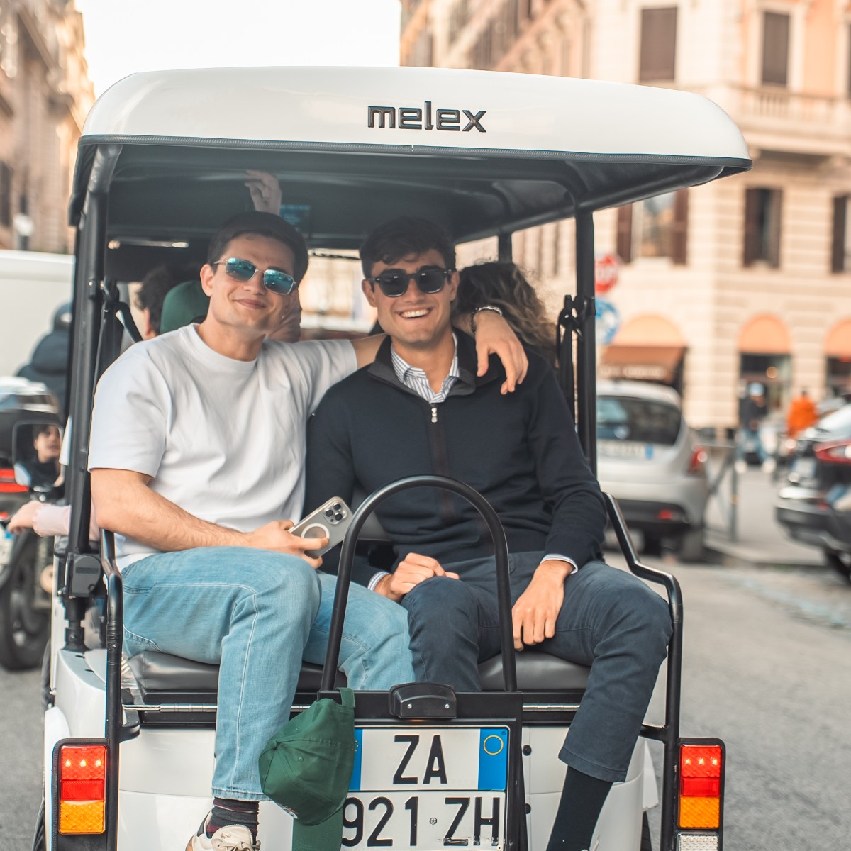 Two people in sunglasses sitting on a cart, smiling on a busy street.