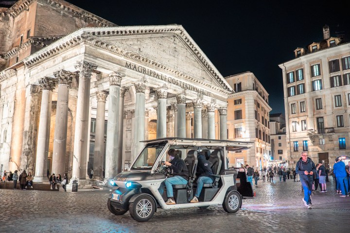 People in a golf cart in front of the Pantheon at night with surrounding buildings illuminated.