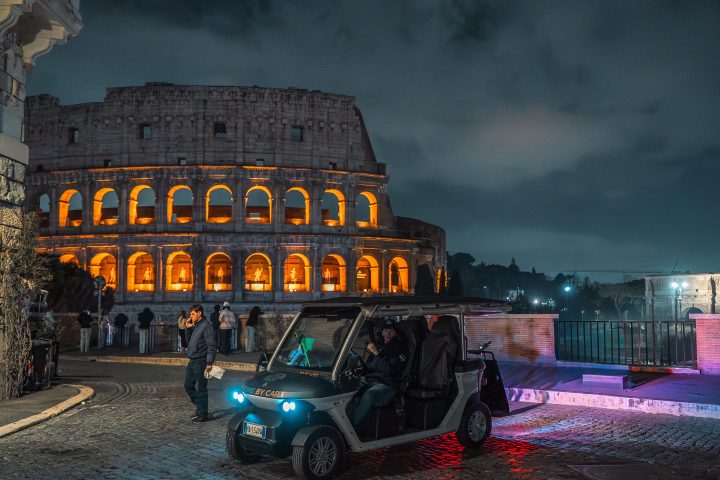 Night view of the illuminated Colosseum with people and a parked golf cart nearby.