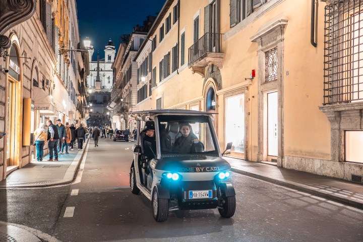 Golf cart driving through a busy European street at night with a church in the background.