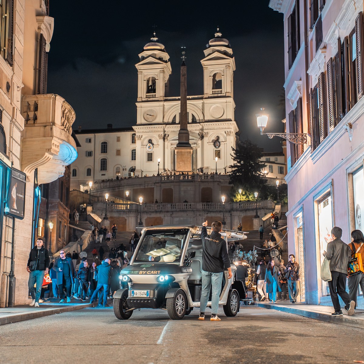 People and a small vehicle on a street near illuminated buildings at night with a large staircase in the background.